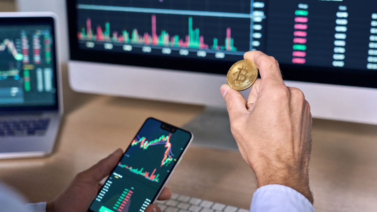 crypto prop trader holding a Bitcoin coin and a mobile phone in front of a monitor with trade charts